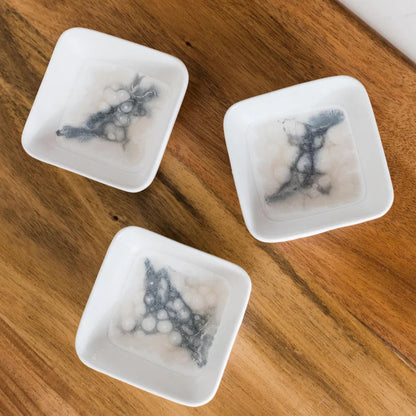 Three square white ceramic dishes with blue resin designs on a wooden surface