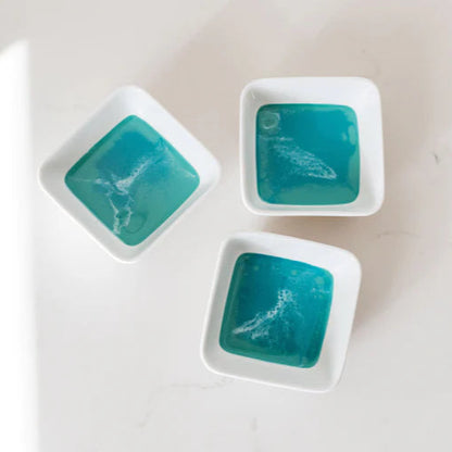 Three small resin white dishes with teal-colored contents on a light gray background