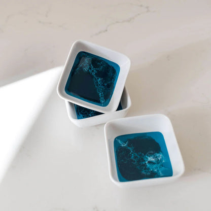 Three resin square white dishes with blue interior patterns on a light surface.