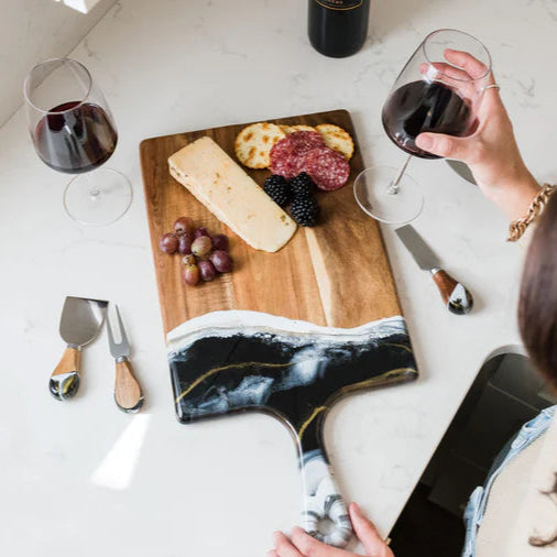 Person holding a glass of red wine with a resin charcuterie board on a marble surface