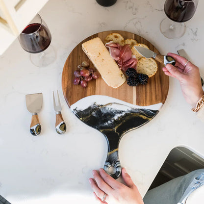 Person preparing a cheeseboard with wine glasses and a laptop on a marble surface