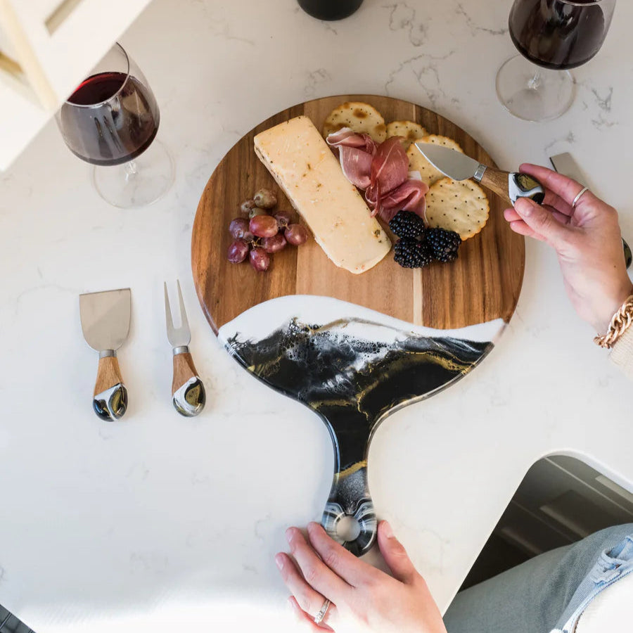 Person preparing a cheeseboard with wine glasses and a laptop on a marble surface