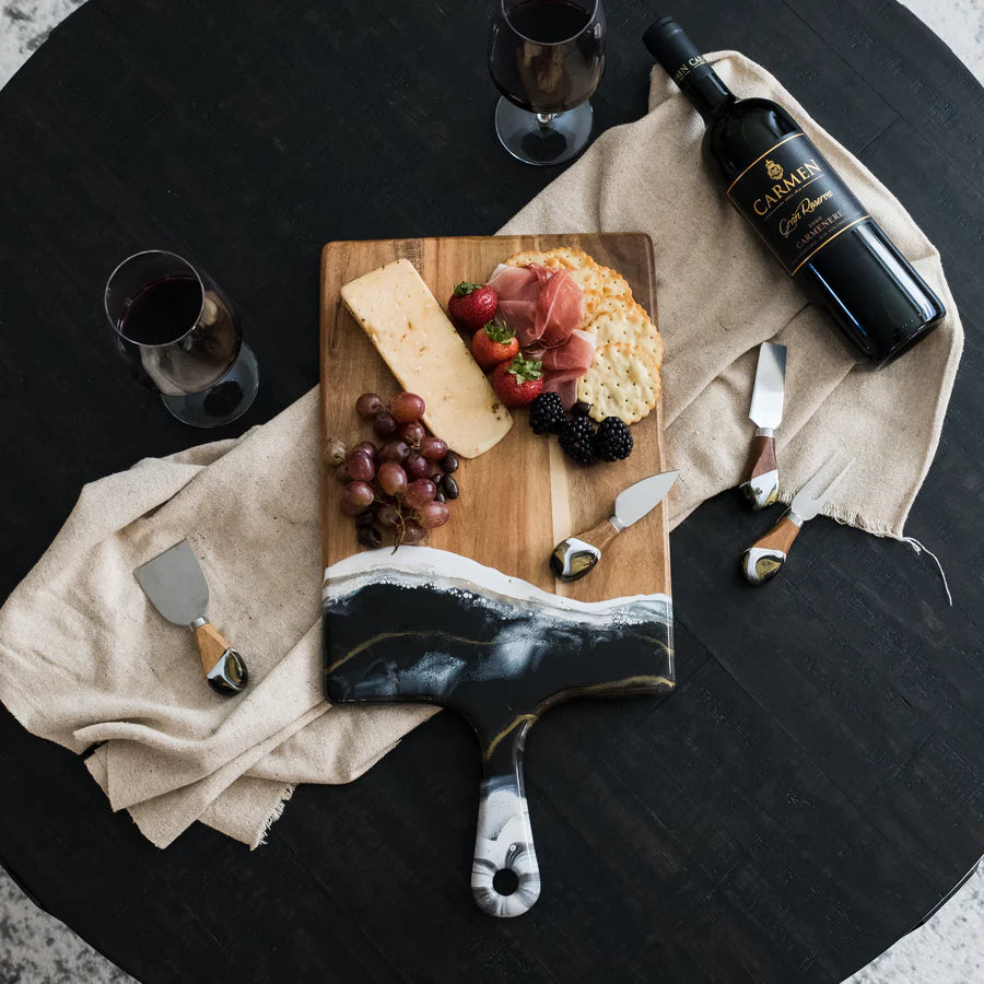 Wooden resin charcuterie board with assorted meats, cheeses, and fruits on a black surface with a bottle of wine and glasses.