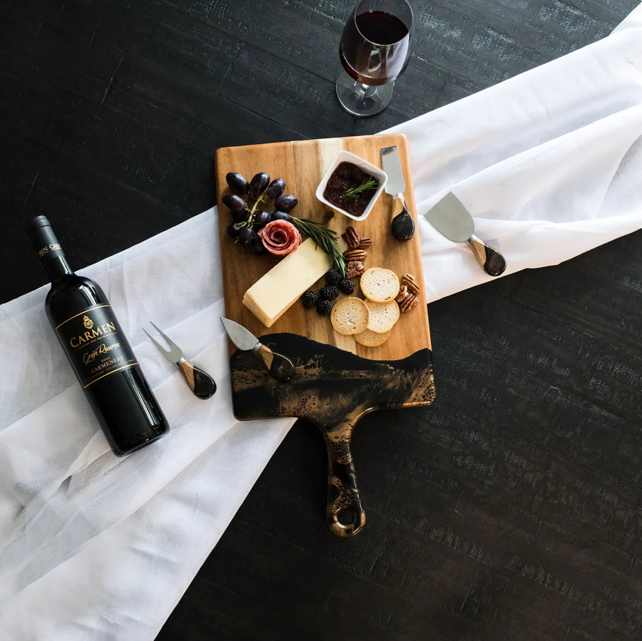 Wine bottle, resin charcuterie board with cheese and grapes, and glass of red wine on a dark surface.