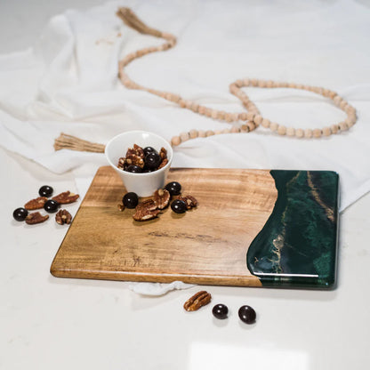 Wooden resin charcuterie board with a bowl of nuts, a green abstract painting, and a necklace on a white fabric background.