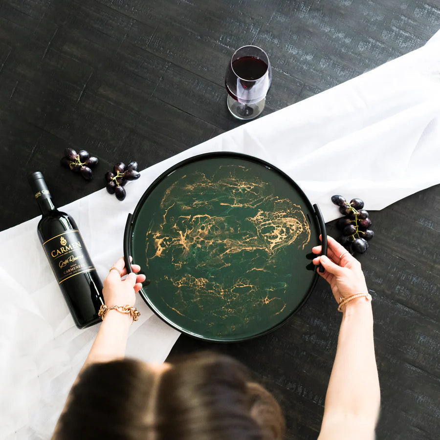 Person holding a decorative resin tray with a bottle of Caramujo red wine and a glass on a table.