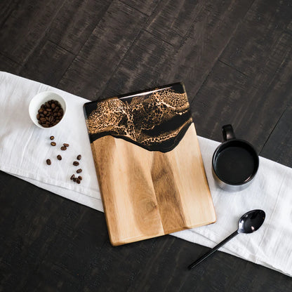 Wooden resin charcuterie board with black resin design on a dark surface with coffee beans and a mug.