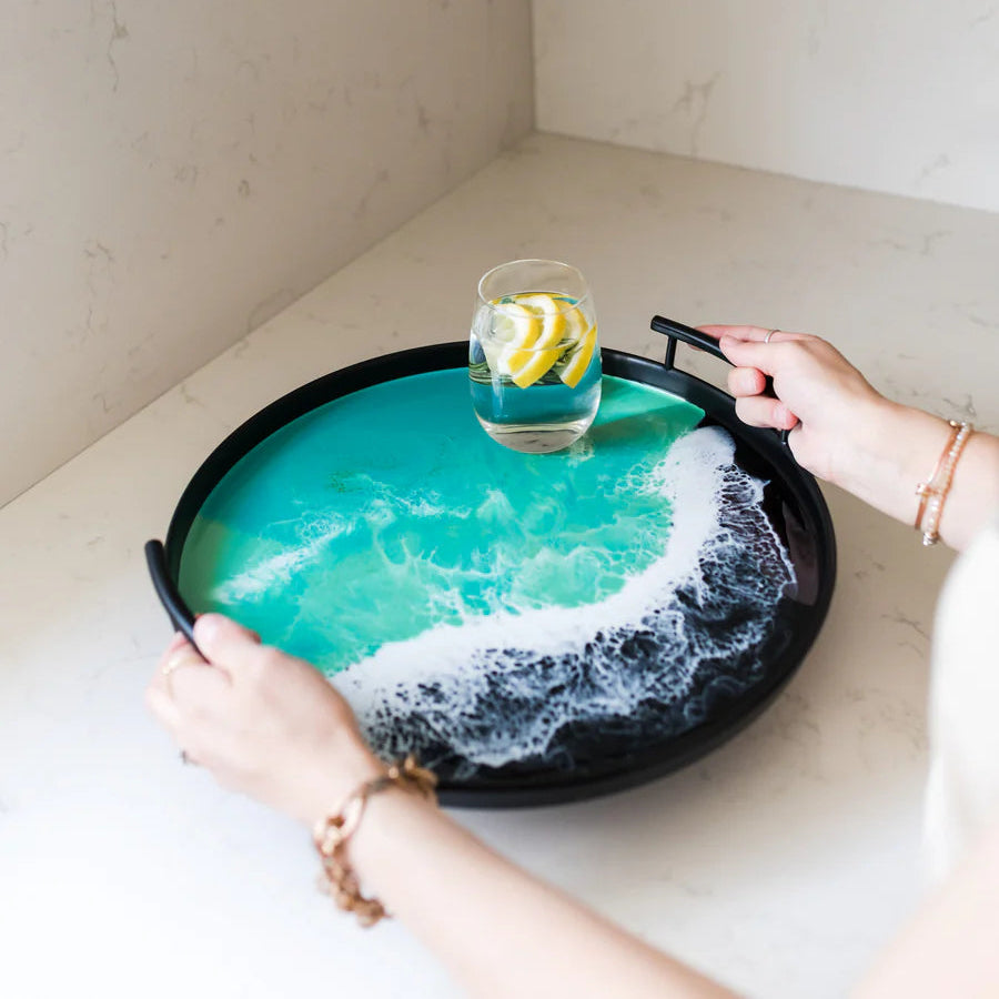 Person holding a decorative resin tray with a glass of water and lemon slices on a kitchen counter.