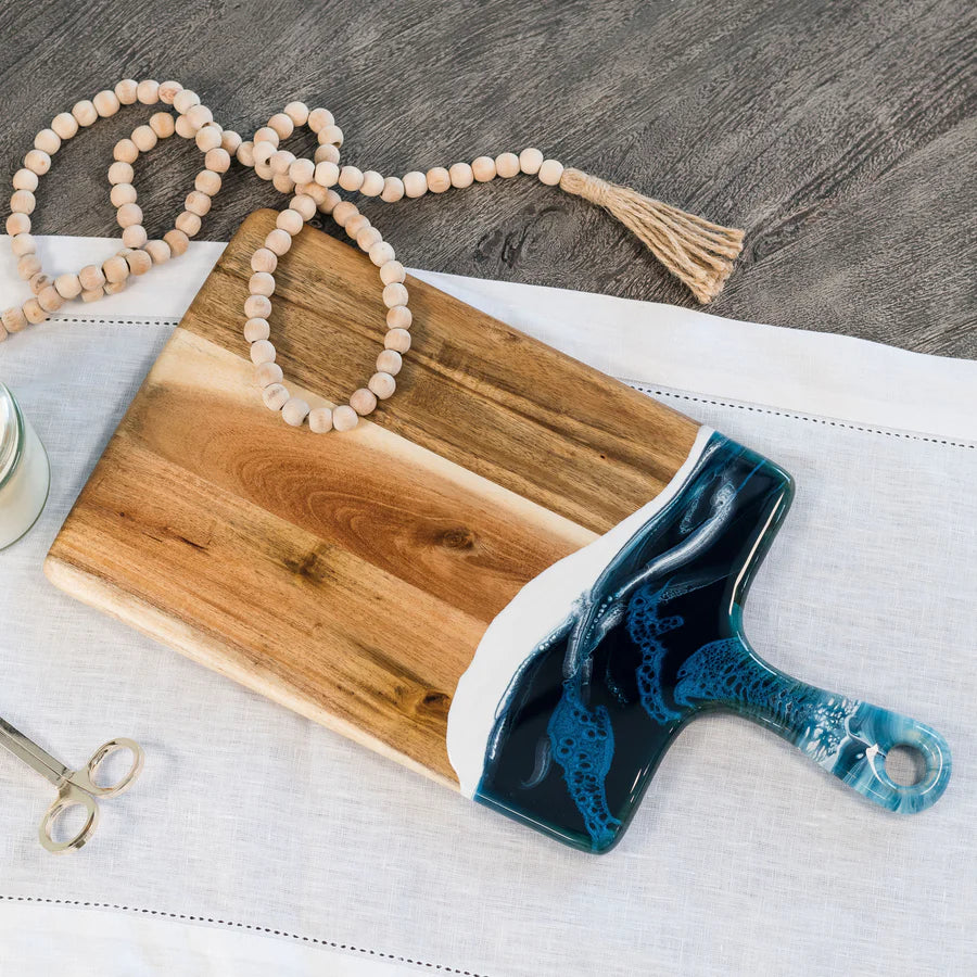 Wooden resin charcuterie board with a blue handle on a white surface, with a beaded necklace in the background.
