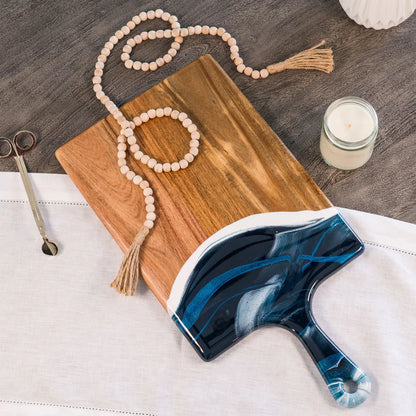 Blue and white marbled paddle brush on a resin charcuterie board with a candle and beads in the background.