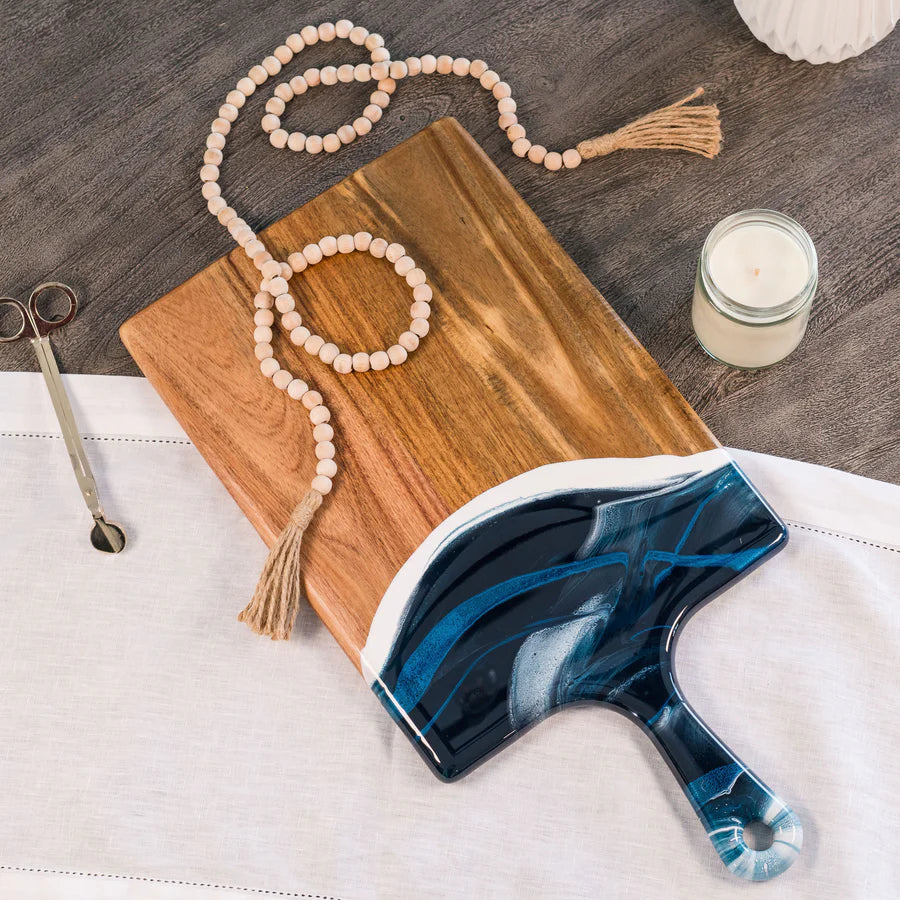 Blue and white marbled paddle brush on a resin charcuterie board with a candle and beads in the background.