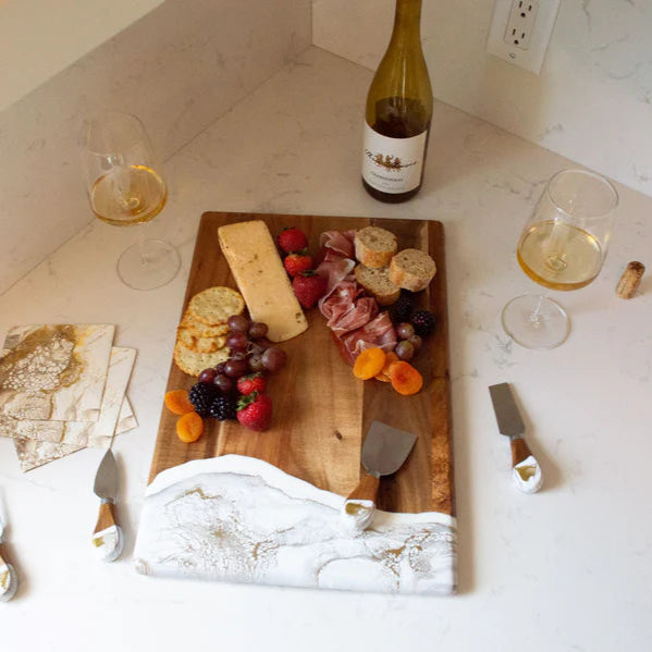 acacia resin serving bread board with cheese and fruit on a marble countertop with wine and glasses.