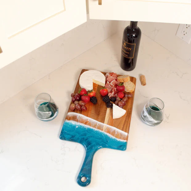 Wooden resin charcuterie board with cheese and fruits on a white surface, accompanied by a bottle of wine and glasses.