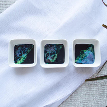 Three small square dishes with colorful resin patterns on a white cloth