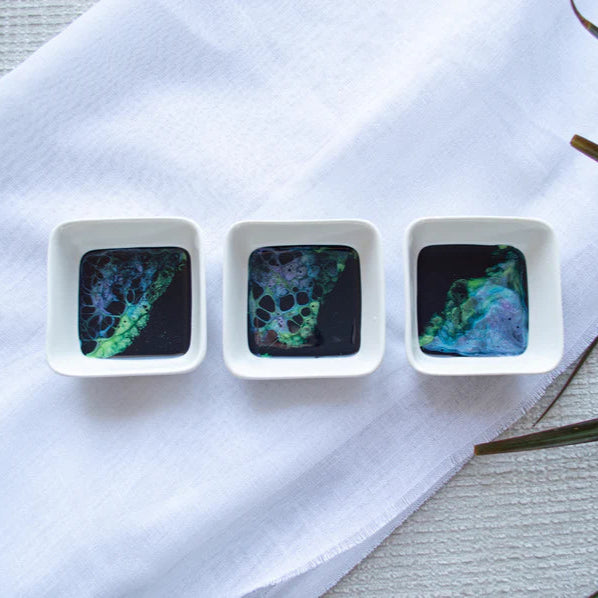 Three small square dishes with colorful resin patterns on a white cloth