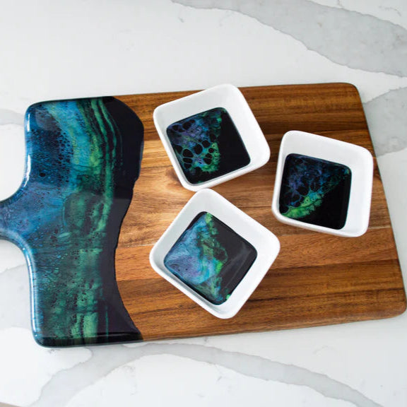 Wooden charcuterie board with a blue and green abstract resin design handle, accompanied by three square dishes with similar abstract patterns on a white surface.