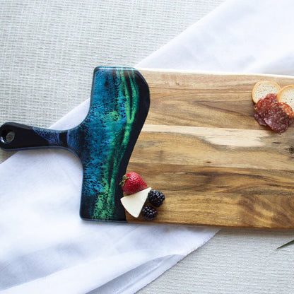 Wooden resin charcuterie board with colorful handle on a white surface with food items.