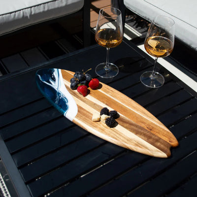 Wooden surfboard-shaped charcuterie board with blue resin accent, holding cheese and fruit on a patio table