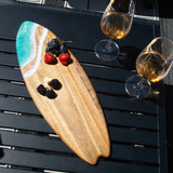 Wooden surfboard-shaped charcuterie board with blue resin accent, holding cheese and fruit on a patio table