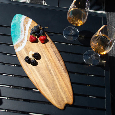 Wooden surfboard-shaped charcuterie board with blue resin accent, holding cheese and fruit on a patio table