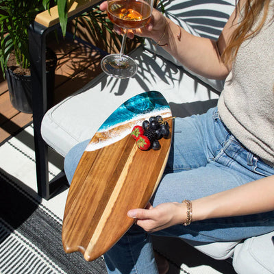 Wooden surfboard-shaped charcuterie board with blue resin accent, holding cheese and fruit on a patio table