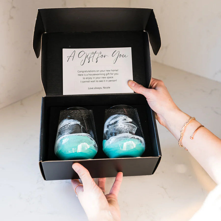 Person holding a black gift box with resin wine glasses and a card inside, set against a kitchen background.