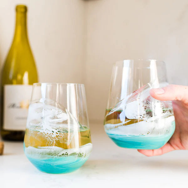 Two stemless resin wine glasses with blue and green liquid, one being held, on a light surface with a blurred bottle in the background.