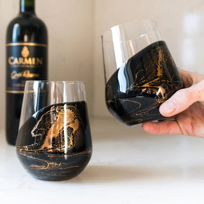 Two stemless resin wine glasses with gold leaf design, one being held, on a white surface with a bottle of Carmel wine in the background.