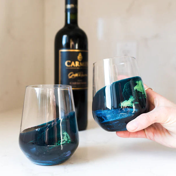 Two wine glasses with blue and green resin designs, one held by a hand, on a white surface with a bottle of Cabsin in the background.
