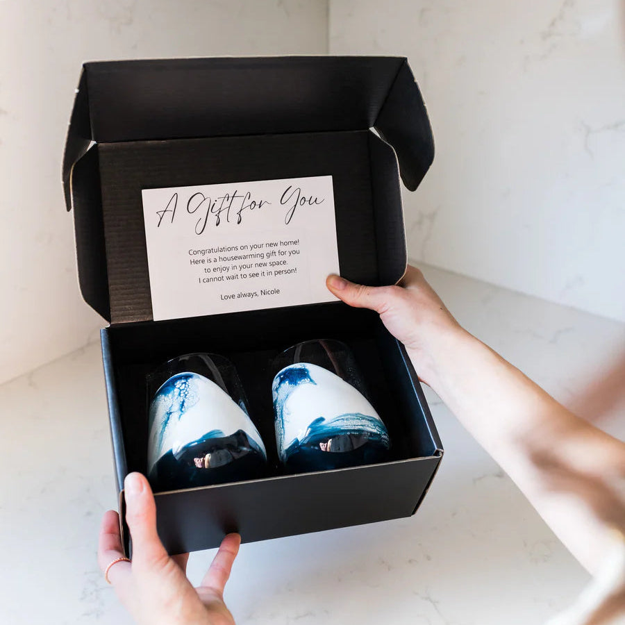 Person holding a black gift box with two resin wine glasses inside, on a light-colored surface.