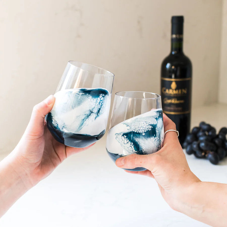 Two hands holding resin wine glasses with blue and white marbled design, bottle of Carmen wine in the background.