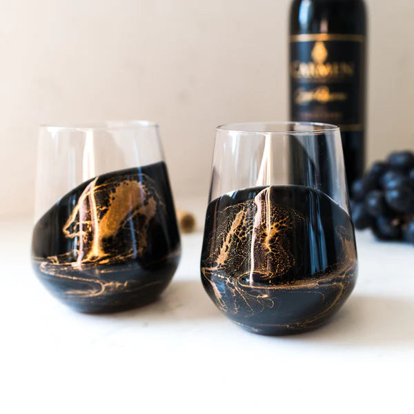 Two stemless resin wine glasses with black and gold design, a bottle of wine, and grapes on a white surface.