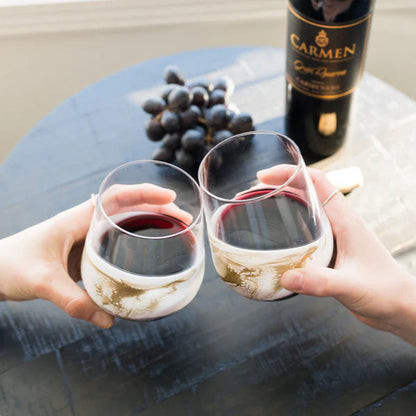 Two people holding resin wine glasses with red wine, a bottle of Carmen, and grapes on a table.
