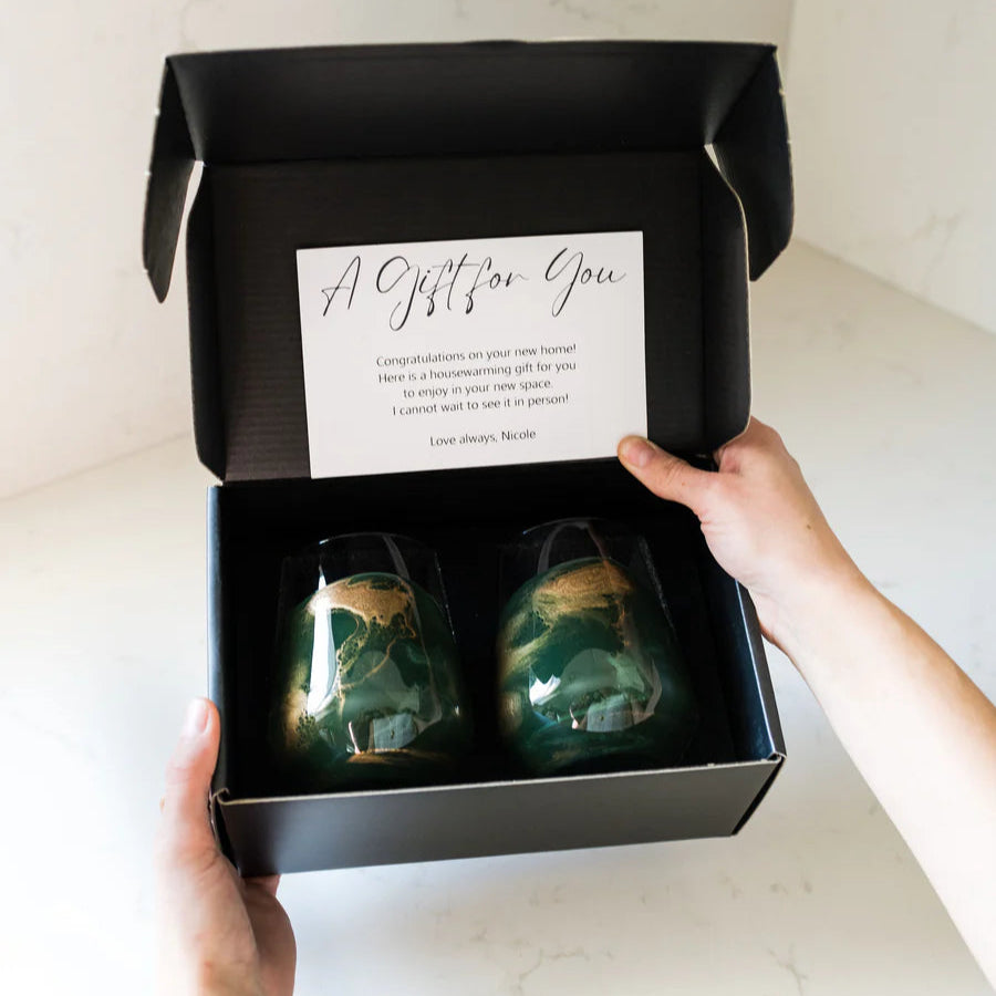 Person holding a black gift box with two green resin wine glasses and a card inside.