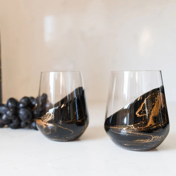 Two stemless resin wine glasses with marbled design next to a bottle of Cabernet Sauvignon and grapes on a light background.