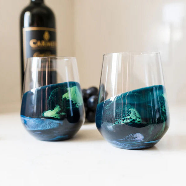 Two stemless wine glasses with blue and green resin designs on a white surface, with a bottle and box in the background.