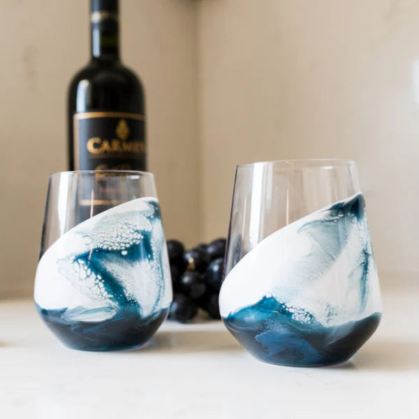 Two resin wine glasses with blue and white marbled design on a table with a bottle of wine and grapes in the background.