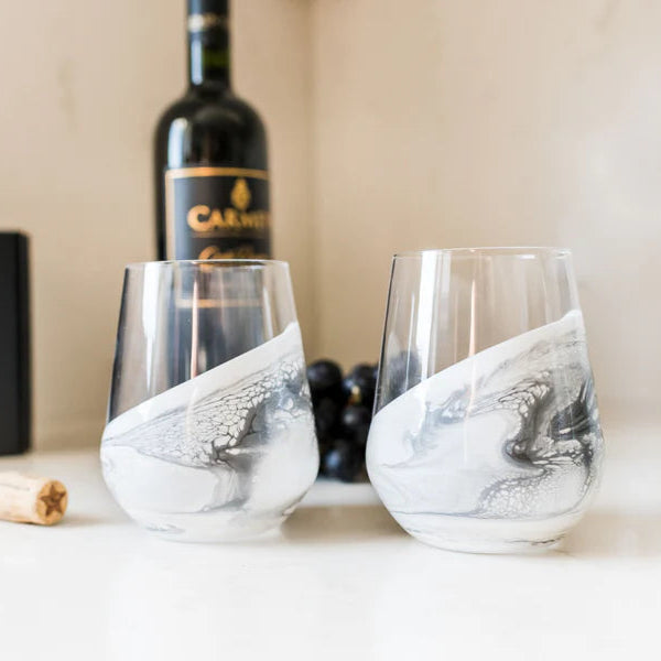 Two marbled resin wine glasses on a table with a bottle of wine and grapes in the background.