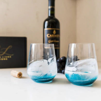 Two blue-tinted resin wine glasses with ice cubes on a table next to a bottle of wine and a box.