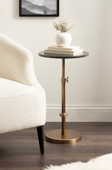 Round dark marble top table with adjustable bronze metal stem and base, holding a white ceramic vase and stacked books beside a cream upholstered chair on wooden floor.