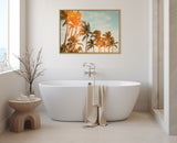 Oval white freestanding bathtub with beige towel draped over side, wooden side table holding bath items and bare branches, and framed palm tree artwork on cream wall.