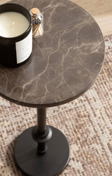 Round dark brown marble table top with light veins, on a slender black metal pedestal base, holding a black candle and a small glass container of matches.