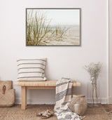 Framed canvas showing beach grass on sandy shore under overcast sky, hanging above a wooden bench with striped pillow and draped striped blanket in a neutral-toned room.
