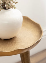 Close-up of textured, matte gold-toned round plant stand top with gently scalloped edges, supporting a white ceramic vase with dried flowers.