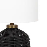 Close-up of a black woven rattan lamp base with a brass accent, topped by a smooth, white fabric lampshade.