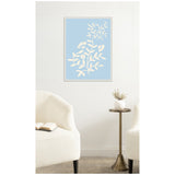 Framed canvas print with cream botanical silhouette on pale blue background, hung above a small round brass side table with a white vase holding dried flowers, flanked by two cream upholstered chairs.
