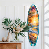 Decorative surfboard wall art with a vivid sunset over ocean waves and palm trees, displayed vertically beside a wooden shelf with green and dried plants in vases.