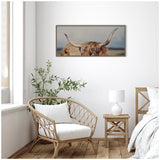 Close-up framed canvas print of a Highland cow with long curved horns and shaggy light brown fur hanging on a white wall above a wicker chair, wooden side table, and lamp.