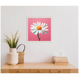 Square framed canvas showing a single white daisy with a yellow center and green stem on a bright pink background, hanging above a wooden table with a vase and stacked books.