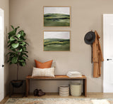 Two framed landscape paintings with green and beige tones hang above a wooden bench holding pillows and books, flanked by a green indoor plant and wall hooks with a hat and coat.